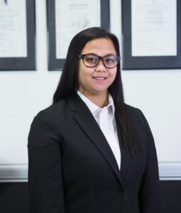 Charisse Castillo, Trainee Practice Manager at Gold Coast Headache and Migraine Clinic, wearing a black blazer and white shirt, standing in front of framed certificates.