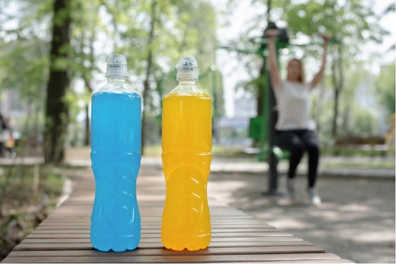 Two brightly coloured drinks filled with electrolytes —blue and yellow—on a park bench, with a person exercising in the background, highlighting the role of hydration and electrolyte intake during physical activity.