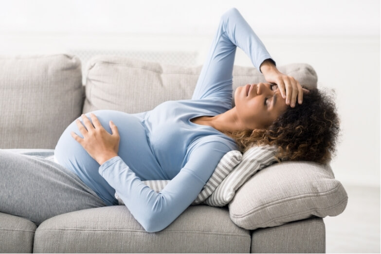 A pregnant woman resting on a couch with her hand on her forehead, visibly experiencing discomfort—illustrating the impact of headaches or migraines during pregnancy.