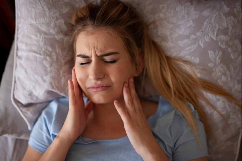 Woman holding her jaw in pain lying in bed — a common sign of TMJ jaw pain and headaches.
